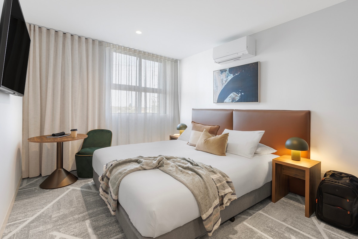 A well-lit sleeping area features a spacious bed with a soft blanket, two bedside tables with lamps, and a comfortable armchair. A small round table is positioned beside a window draped with light curtains, while a TV is mounted on the wall opposite the bed.