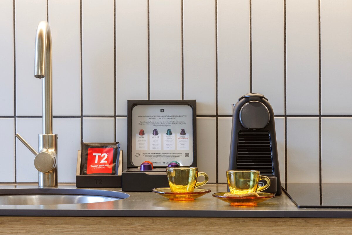 A modern kitchen countertop is featured, displaying a sleek coffee machine, two amber-colored cups, and a box of assorted coffee pods. A tea package is visible beside the coffee setup, and a mounted card outlines beverage options.