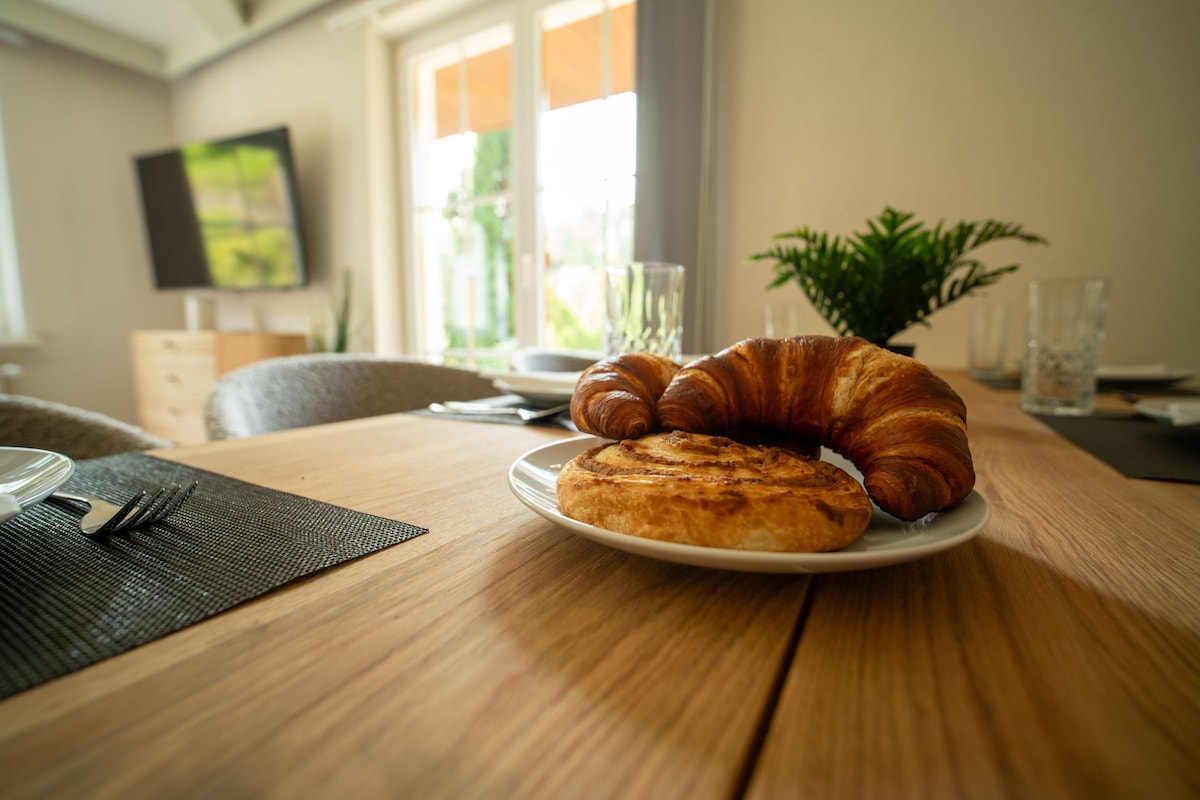 A wooden dining table is set with a plate of freshly baked pastries, including a croissant and a spiraled sweet roll. Clear glassware and cutlery are arranged neatly nearby, while a green plant adds a touch of nature to the inviting dining area.