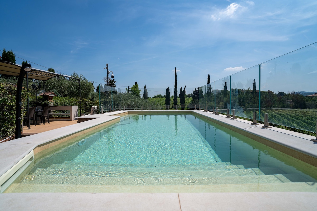 A serene salt-water pool is set against a backdrop of lush vineyards and tall cypress trees. Glass fencing offers unobstructed views of the landscape, while a shaded dining area nearby invites outdoor meals.