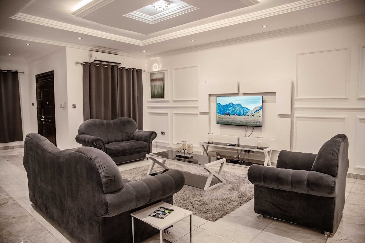 A modern living area is presented with plush gray sofas arranged around a sleek glass coffee table. A large television is mounted on the wall, displaying a landscape image. Soft lighting complements the neutral tones, while curtains frame the windows.