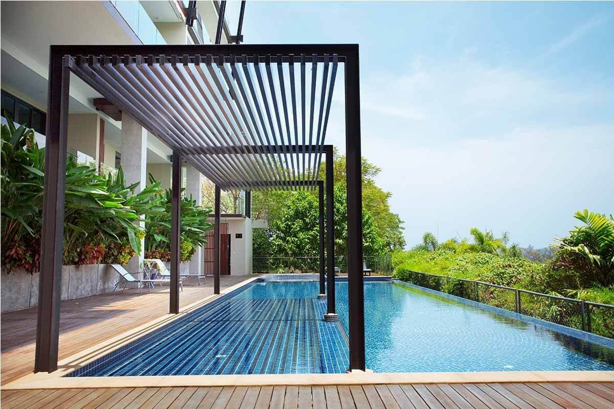 An outdoor swimming pool is framed by sleek, modern architecture. Lush greenery surrounds the pool area, complemented by a shaded seating structure with horizontal slats that provides a tranquil space to relax. The water reflects sunshine, inviting a refreshing dip.
