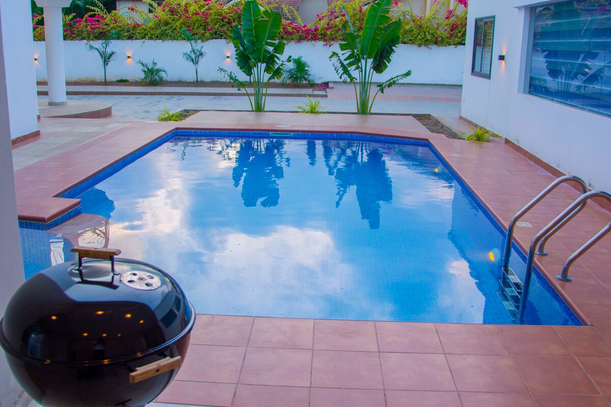 A clean, inviting swimming pool is framed by smooth tiled surfaces and lush greenery. A stylish grill is positioned nearby, complementing the outdoor space. The area is illuminated softly, creating a relaxing ambiance with reflections shimmering on the water's surface.