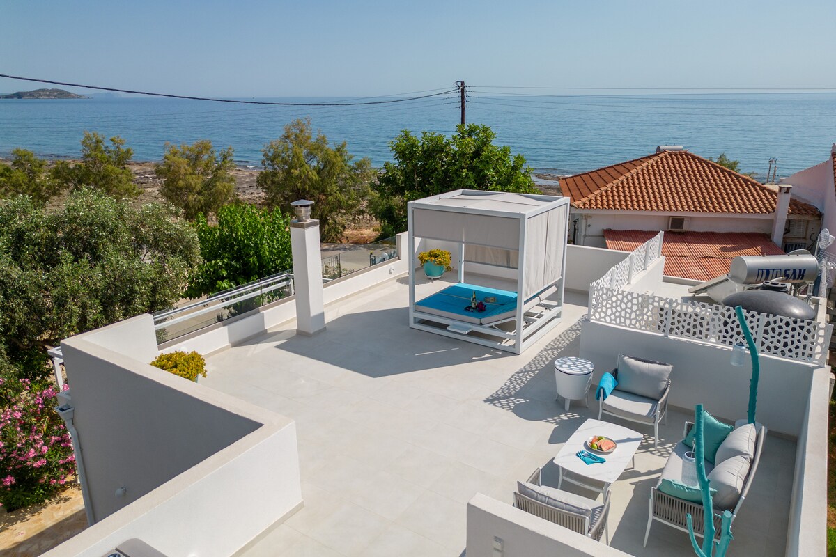 A spacious rooftop terrace overlooks the sea, featuring a large, shaded lounge area with comfortable furnishings. A secluded cabana invites relaxation, while vibrant greenery and coastal views enhance the serene environment.