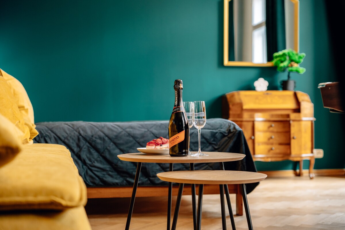 A modern sleeping area features a spacious bed with yellow and dark bedding. A set of two round tables holds a bottle of champagne and glasses, alongside a plate with pastries. A vintage wooden dresser and a large mirror are visible on the wall.