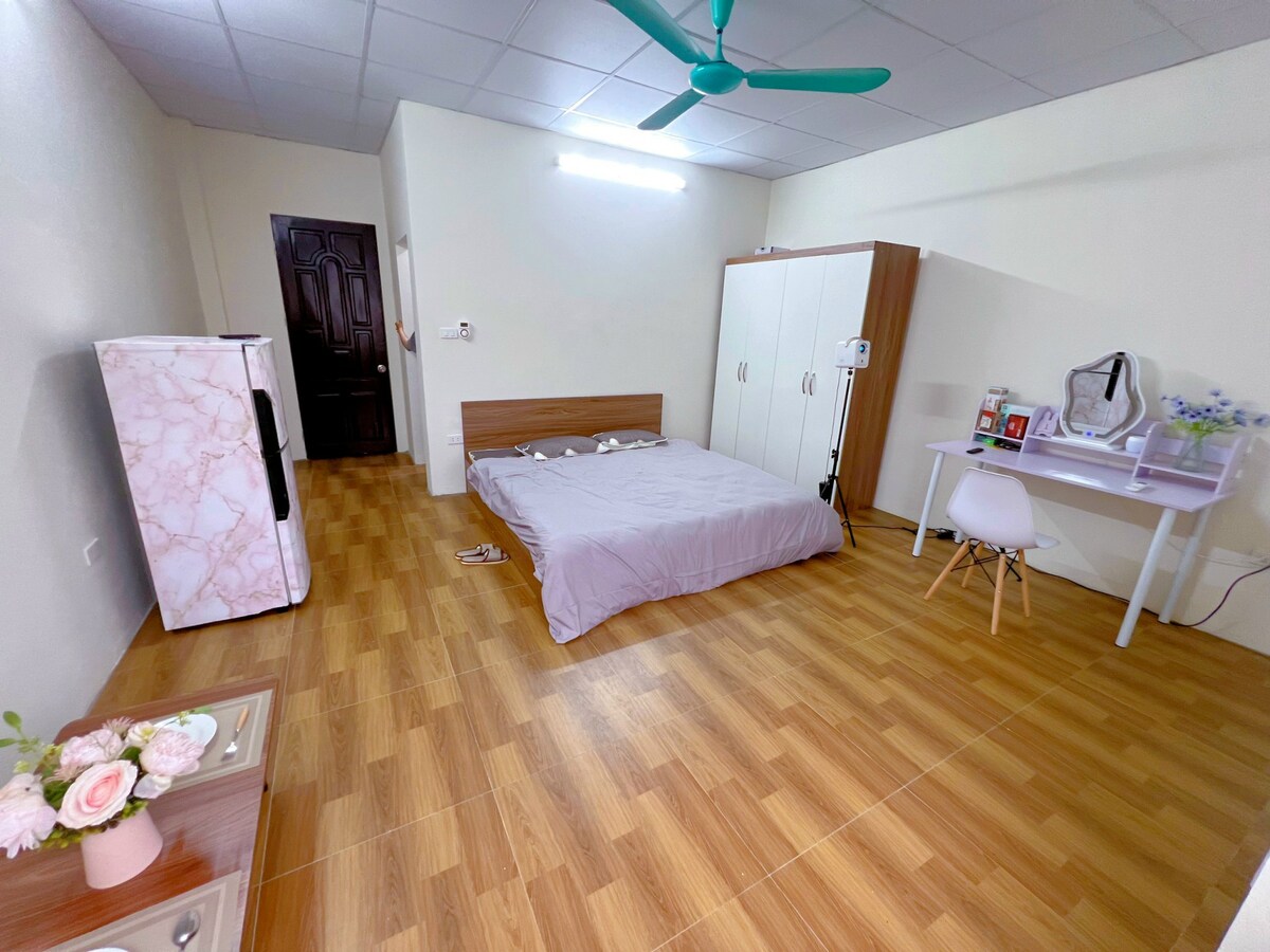 A cozy studio apartment is shown, featuring a spacious bed with a soft cover. A minimalist desk with a chair is placed near the wall, while a large wardrobe stands adjacent. A marble-patterned refrigerator is visible, complementing the warm wood flooring.