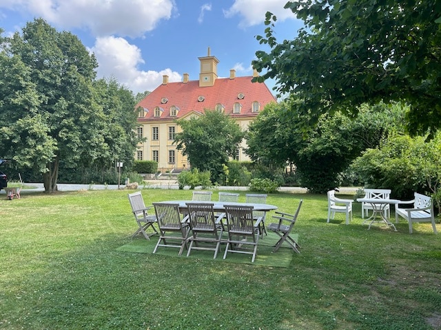 A spacious garden area features multiple seating arrangements, including a large table surrounded by wooden chairs. The lush green grass is complemented by trees offering shade, with a charming yellow building visible in the background under a partly cloudy sky.