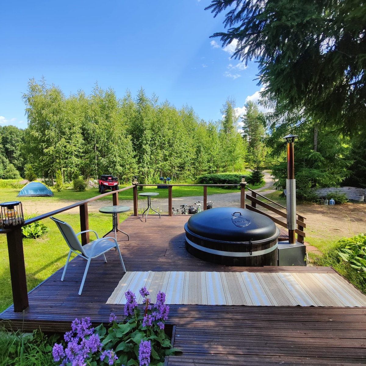 A wooden terrace is visible, featuring a round hot tub and a small table with a chair. Surrounding greenery includes trees and a flower bed with purple blooms. The scene is complemented by a distant view of a grassy area and a parked vehicle.