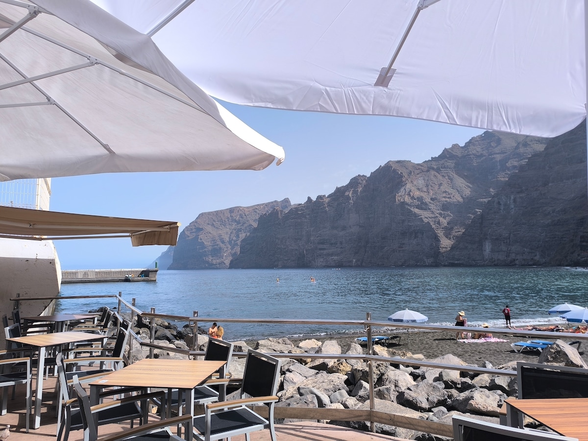An outdoor dining area is featured with several wooden tables and chairs set on a deck overlooking the ocean. The cliffs of Los Gigantes rise majestically in the background, while umbrellas provide shade for guests enjoying the view.