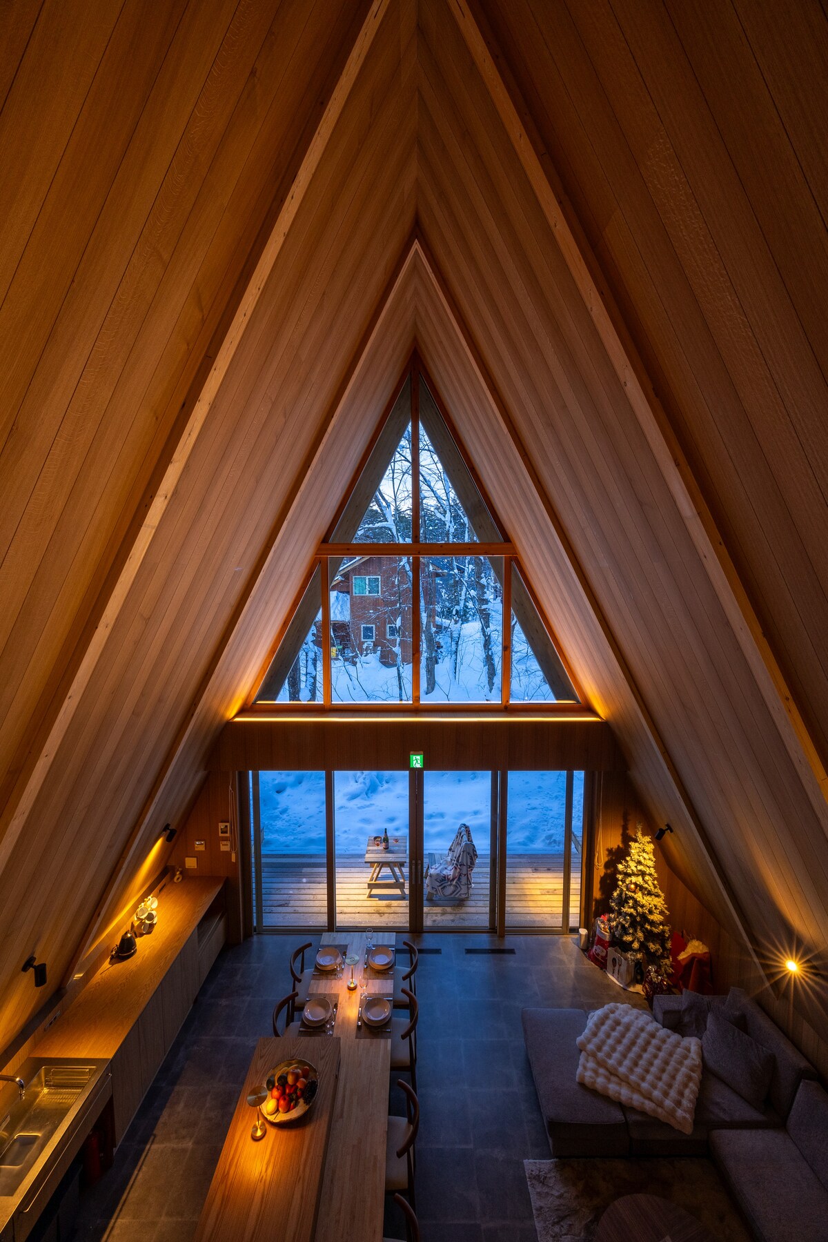 The A-frame design showcases tall wooden walls meeting at a peak, with large windows offering views of the snowy landscape outside. A long dining table is positioned beside a cozy seating area, illuminated by soft lighting, while a festive tree adds a touch of warmth.
