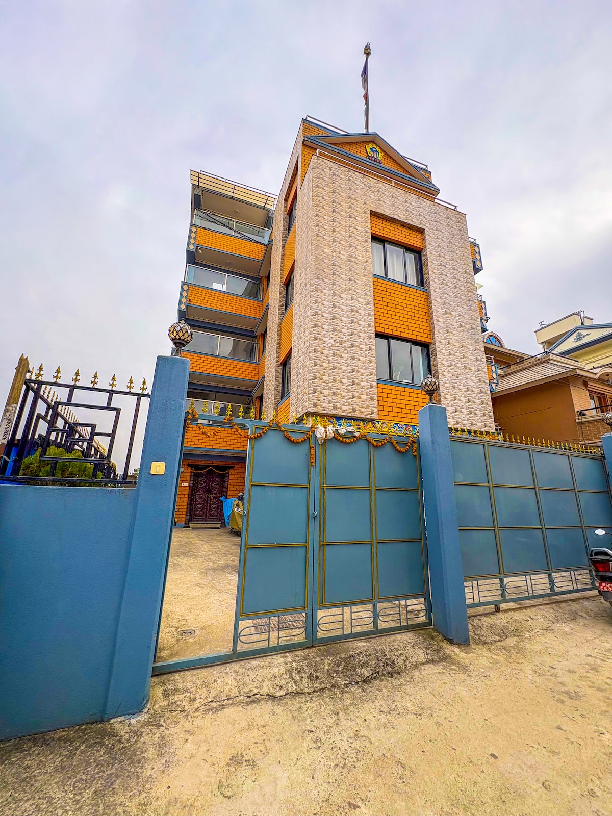 The exterior of a multi-story building is showcased, featuring a mixture of light brick and orange tiles. A gated entrance with decorative elements stands in front, providing access to the property. The structure is three levels high, with multiple windows allowing natural light.