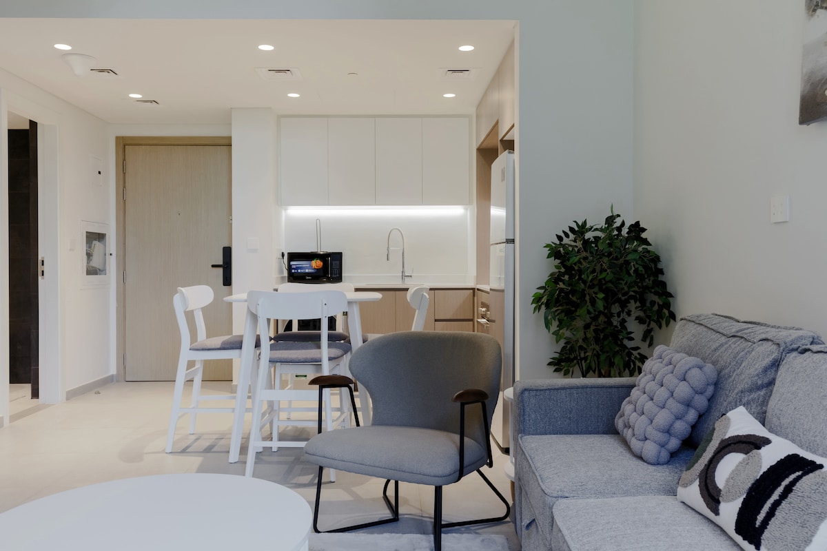 A modern living area features a gray sofa alongside a contemporary armchair, with a round coffee table in front. A dining table with high chairs is visible, set against an open kitchen that includes sleek cabinetry and essential appliances. Natural light brightens the space.