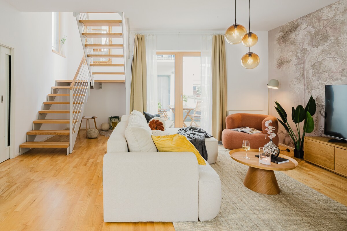 A spacious living area is shown, featuring a soft sofa and a cozy armchair. Large glass doors open onto a patio, allowing natural light to flow in. A wooden coffee table is positioned centrally, complemented by decorative elements. A staircase leads to the upper floor.
