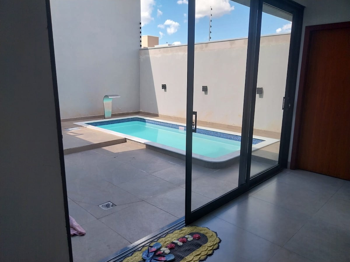 A private pool area is visible through large glass doors, featuring a turquoise pool framed by light-colored tiles. The surrounding walls are finished in neutral tones, enhancing the outdoor space. Natural light illuminates the area, with clouds reflecting in the sky above.