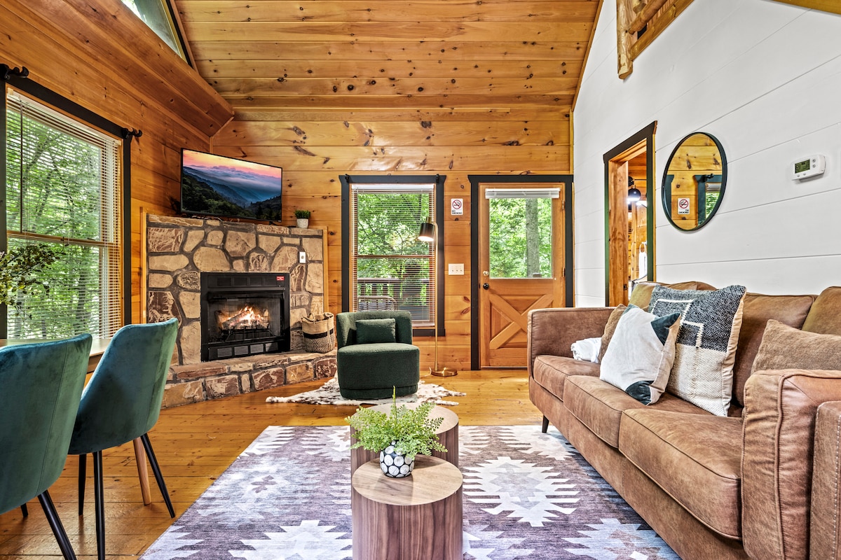 The living area features warm wooden walls and vaulted ceilings, with a cozy stone fireplace as the focal point. Comfortable seating is provided by a brown sofa and green chairs. A large window offers views of the surrounding nature, while a TV hangs above the fireplace.