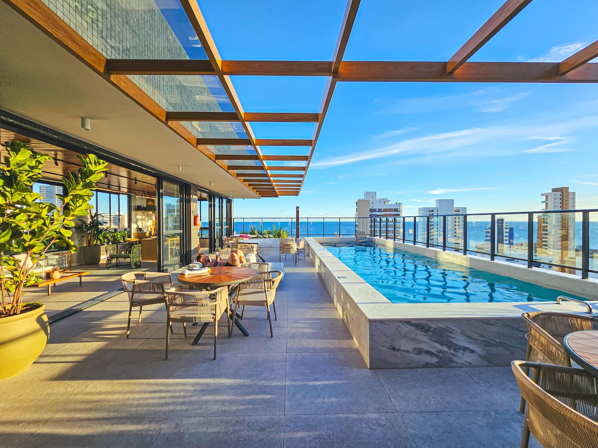 An outdoor terrace showcases a swimming pool bordered by modern stonework, complemented by a spacious dining area. Panoramic views of the skyline and ocean can be seen in the background. The space features a wooden pergola overhead, providing structure and shade.