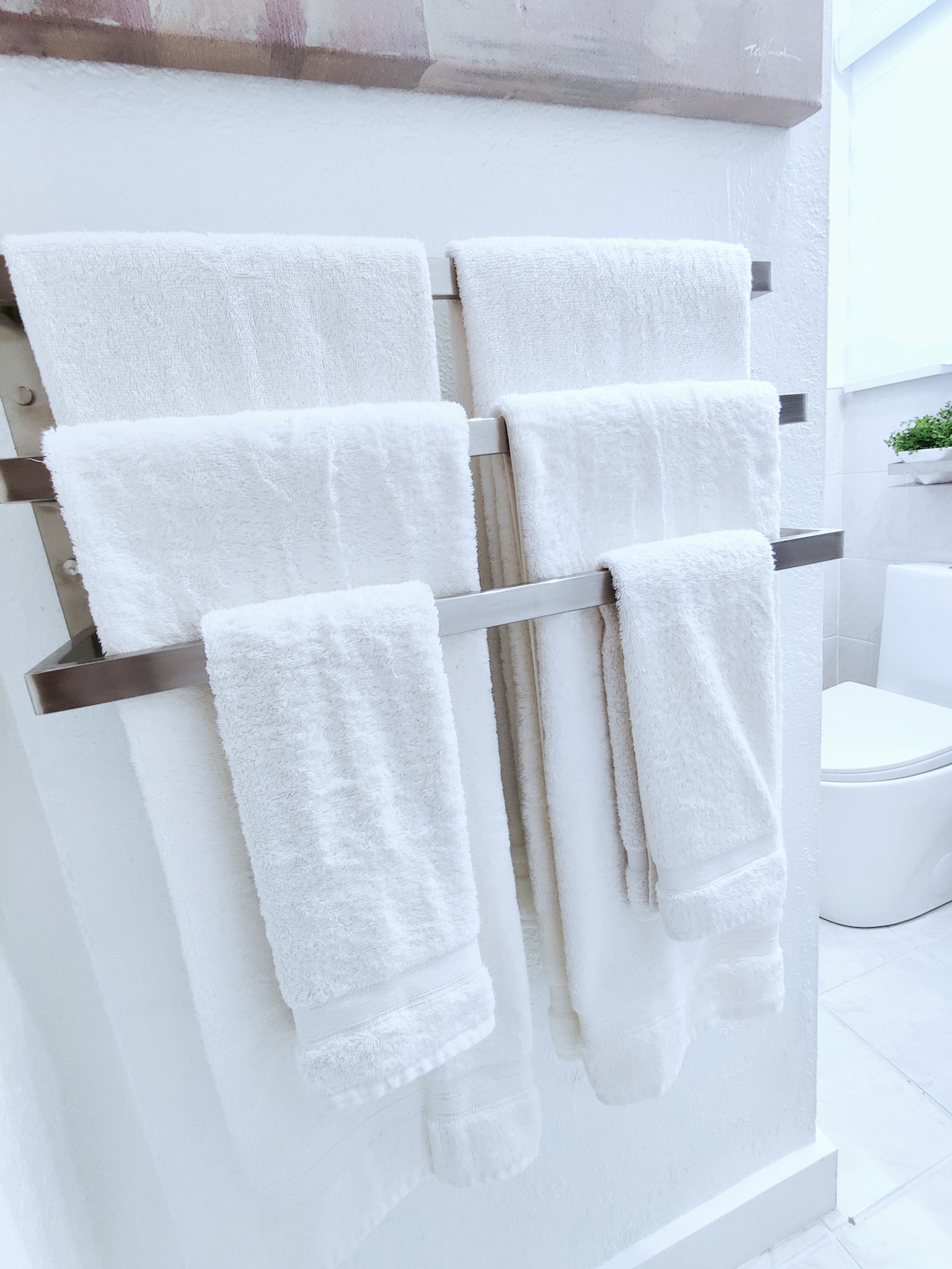 Four neatly arranged white towels hang on a modern towel rack against a clean, light-colored wall. A glimpse of a toilet is visible in the background, contributing to the bright and uncluttered bathroom atmosphere.