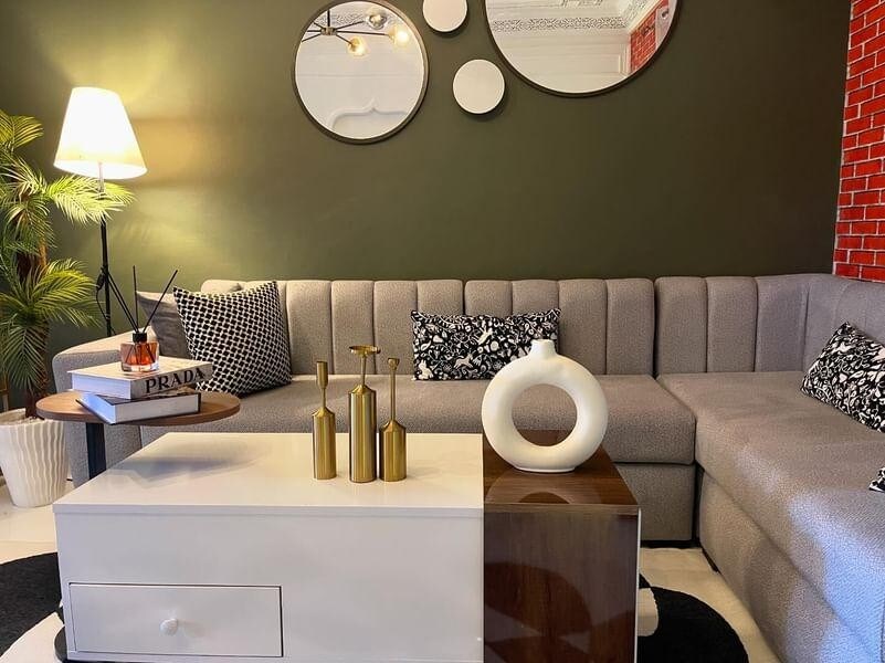 A modern living area is featured with a cozy sectional sofa in neutral tones, complemented by black and white patterned pillows. A round, decorative coffee table sits in front, adorned with a few sleek vases and a stylish magazine. Warm lighting enhances the inviting ambiance.