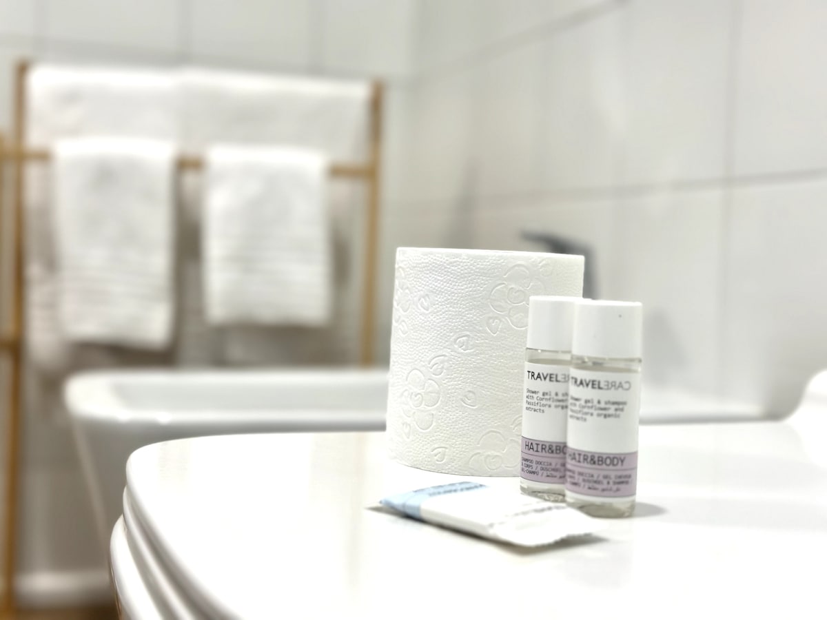 A clean bathroom countertop is presented, featuring travel-sized toiletries for hair and body care. A roll of toilet paper sits alongside the toiletries. In the background, neatly arranged towels are visible on a wooden rack, contributing to the overall functionality of the space.