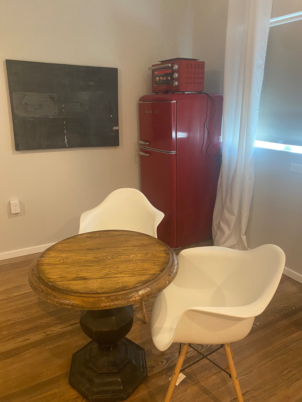 A small dining area features a round wooden table with a pedestal base and two white chairs. A bright red refrigerator stands nearby against a light-colored wall, while a piece of abstract art hangs above the table, complementing the warm wood flooring.
