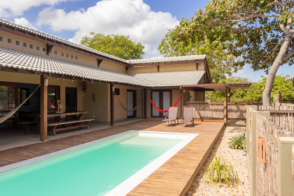 The outdoor area features a private pool surrounded by a wooden deck, complemented by two lounge chairs. Hammocks are strung between posts, and the building's overhang provides shaded seating space. Lush greenery and a gravel border enhance the inviting environment.