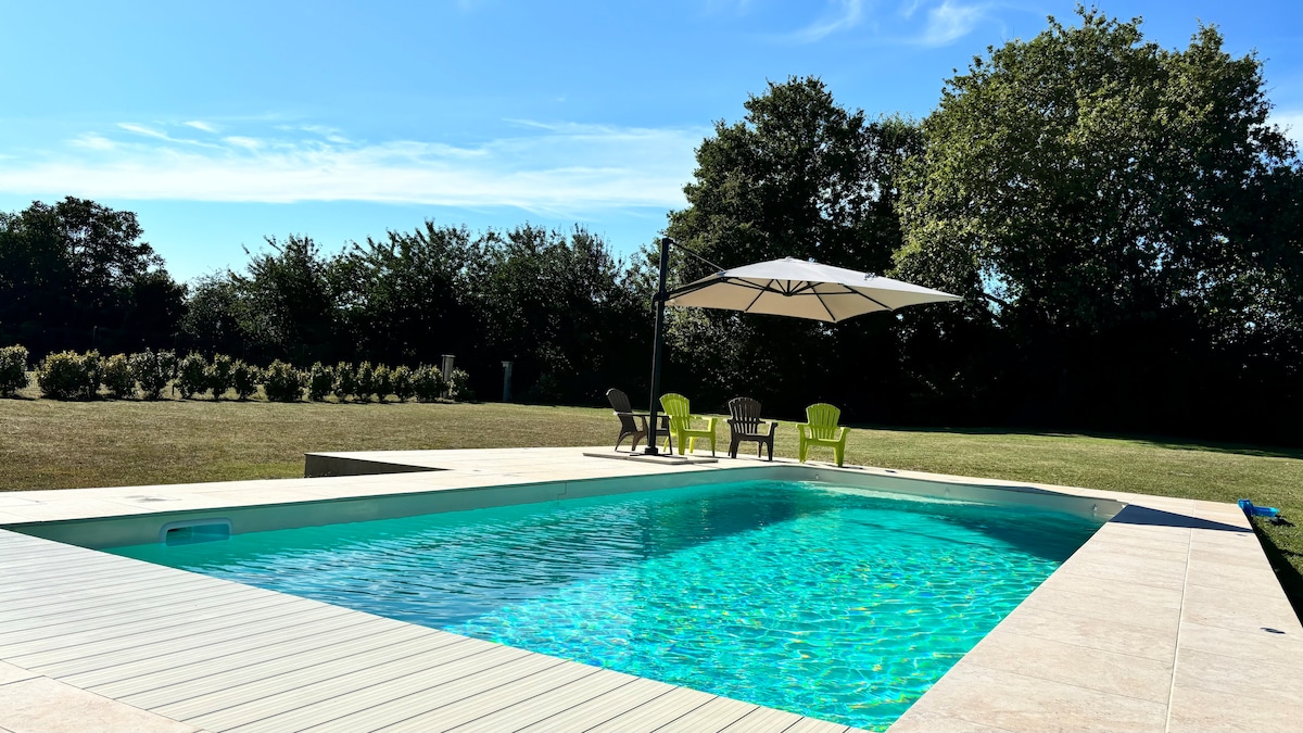 An outdoor swimming pool is framed by a spacious deck and surrounded by a green landscape. Comfortable lounge chairs are positioned under a large umbrella, providing a shaded area. The clear blue water of the pool reflects the sunny sky, creating a serene atmosphere.