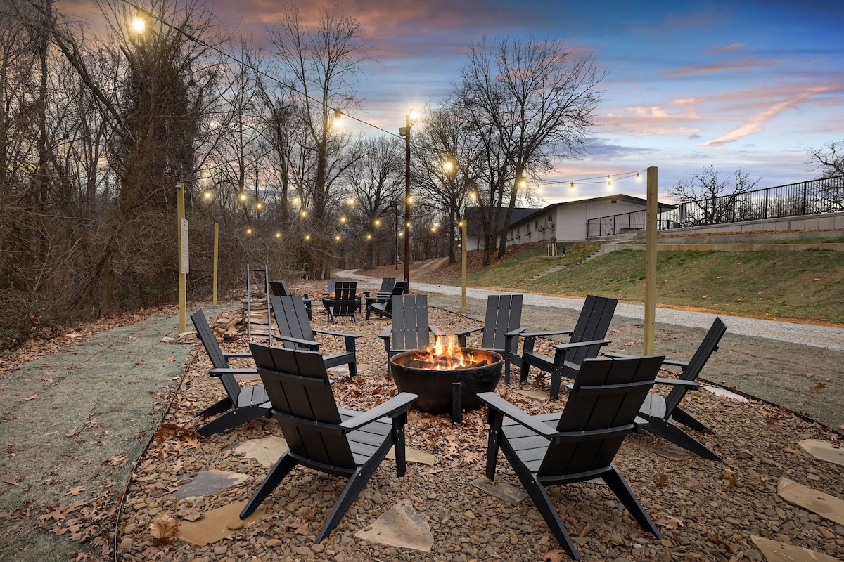 An outdoor seating area features multiple black chairs arranged around a central fire pit. Warm light from string lights above enhances the cozy ambiance, while bare trees line the background against a twilight sky, creating an inviting space for gatherings.