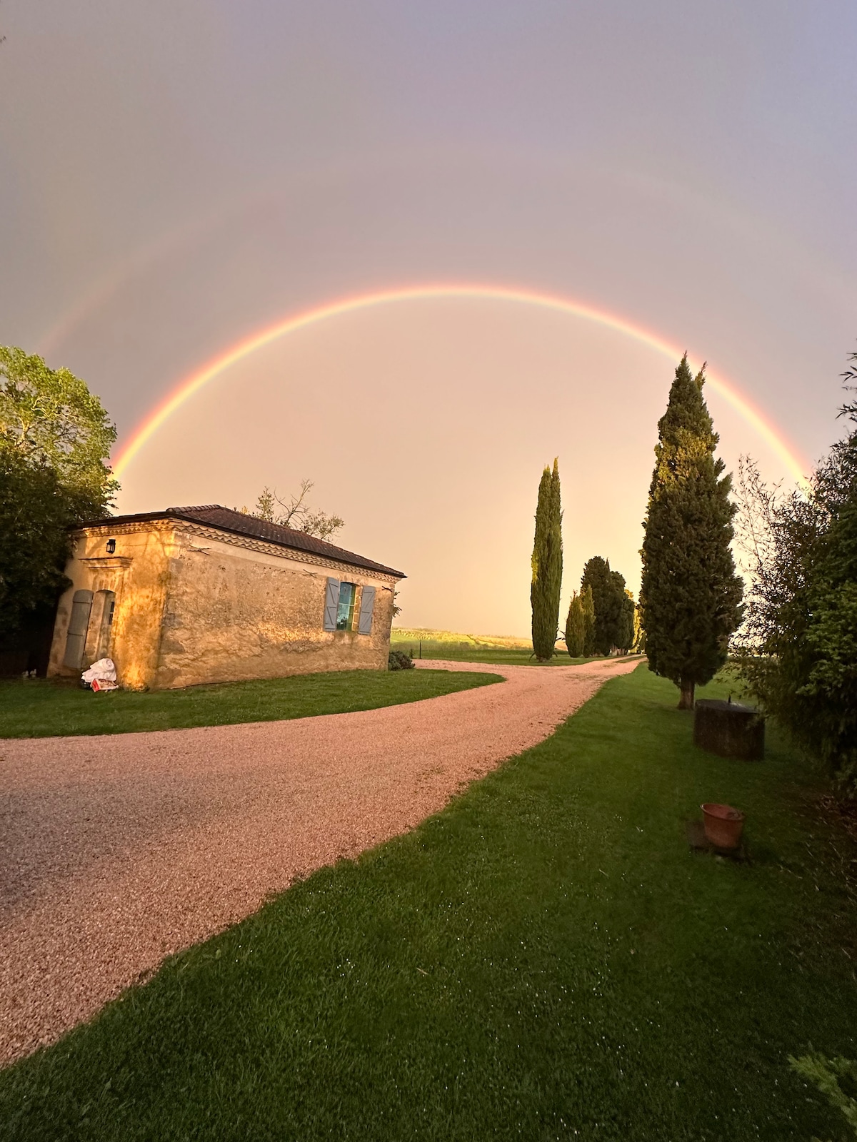 A charming building is positioned along a gravel pathway flanked by lush greenery. Towering cypress trees rise nearby, and an impressive rainbow arches across the sky, creating a serene atmosphere. The structure features blue shutters, reflecting natural light from the setting sun.