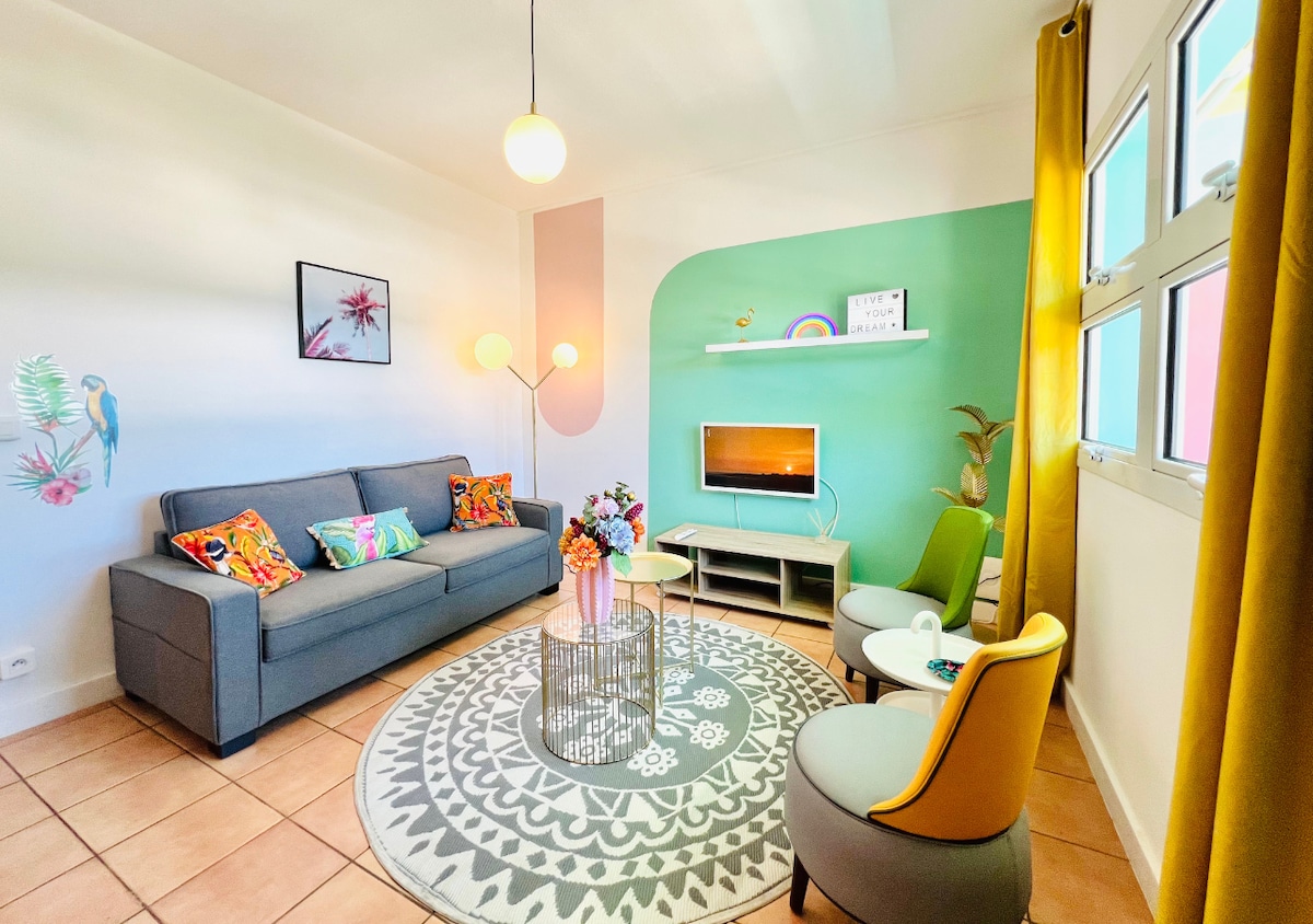 The living room features a gray sofa adorned with colorful cushions, paired with two accent chairs. A round coffee table sits on a patterned rug, and a flat-screen television is mounted on the wall. Large windows allow natural light to brighten the space.
