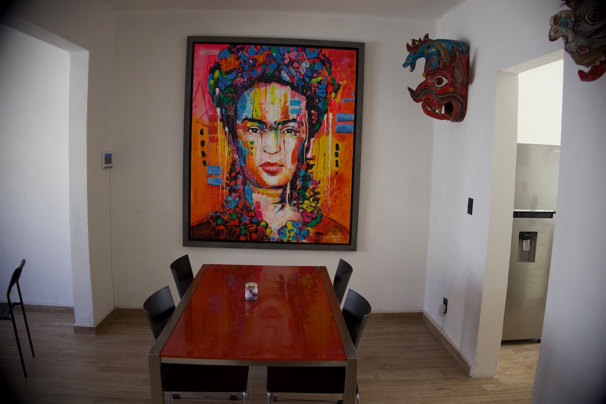 A dining area features a rectangular table surrounded by black chairs. A large, colorful artwork adorns the wall, creating a vibrant focal point. Unique masks are displayed in the corners, adding character to the space, which is illuminated by natural light.