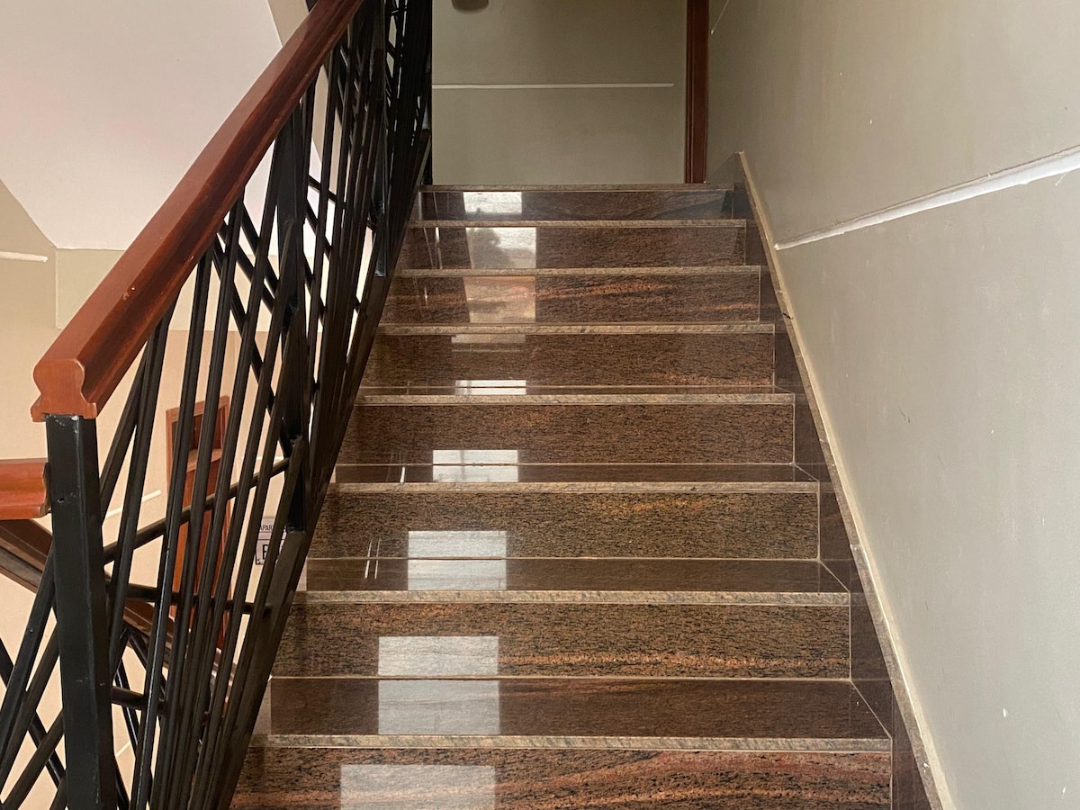 A well-maintained staircase features polished granite steps and a sturdy wooden railing. Natural light is reflected off the surface, enhancing the cleanliness of the space. The walls are painted in a neutral tone, contributing to a modern and inviting appearance.