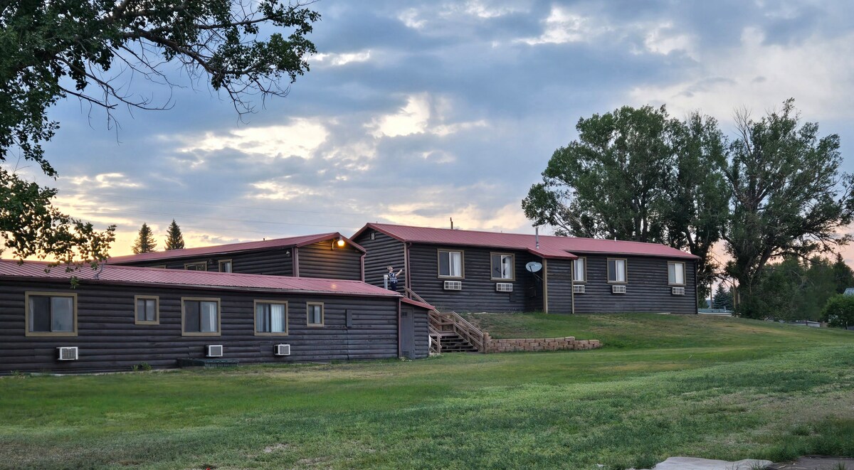 The exterior of the motel features wooden siding, a red metal roof, and air conditioning units. A grassy area surrounds the building, and a set of steps leads to the entrance. Soft clouds are visible in the sky during dusk, creating a serene backdrop.