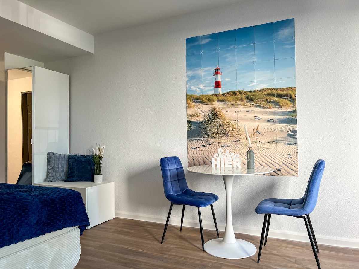 A bright studio apartment features a cozy sleeping area with a patterned blue blanket and a decorative wall art of a coastal scene. A white table is accompanied by two blue chairs, creating a small dining area, while a plant adds a touch of greenery.