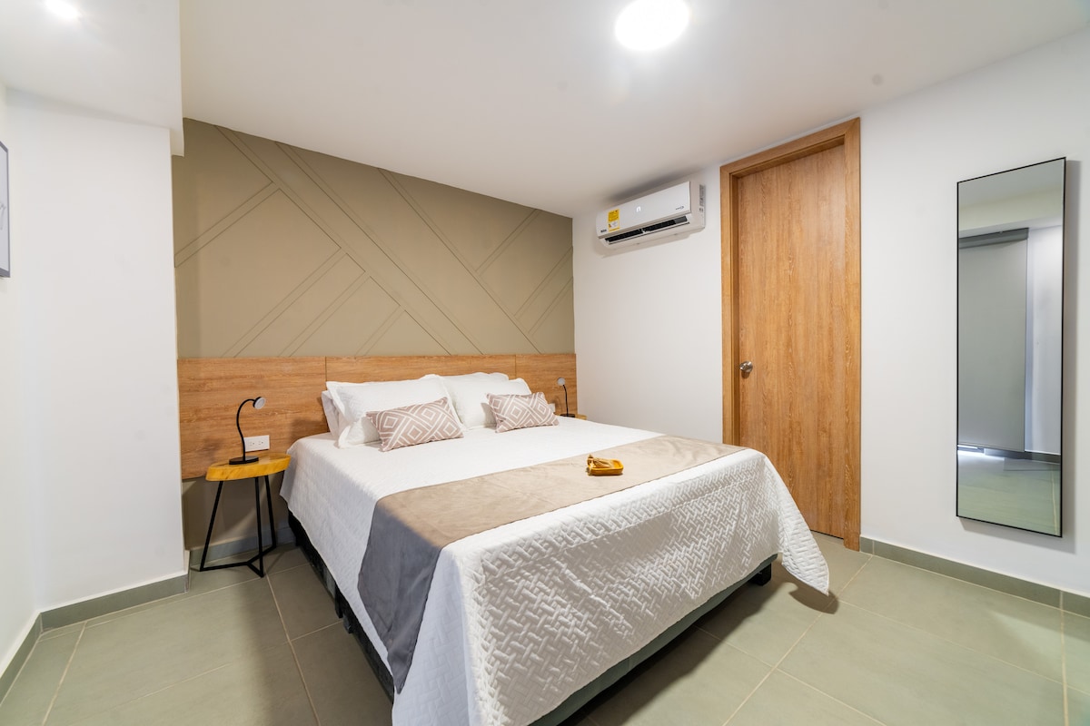 A well-lit room features a spacious bed with neutral bedding against a modern accent wall. A small bedside table holds a decorative item. A mirror and air conditioning unit are visible, enhancing the room's functionality and comfort.