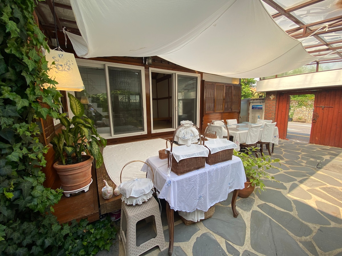 An outdoor seating area features a table covered with a white tablecloth, surrounded by woven chairs. Lush greenery and potted plants enhance the natural setting. Large windows provide views into the interior space, while a canopy offers shade over the area.