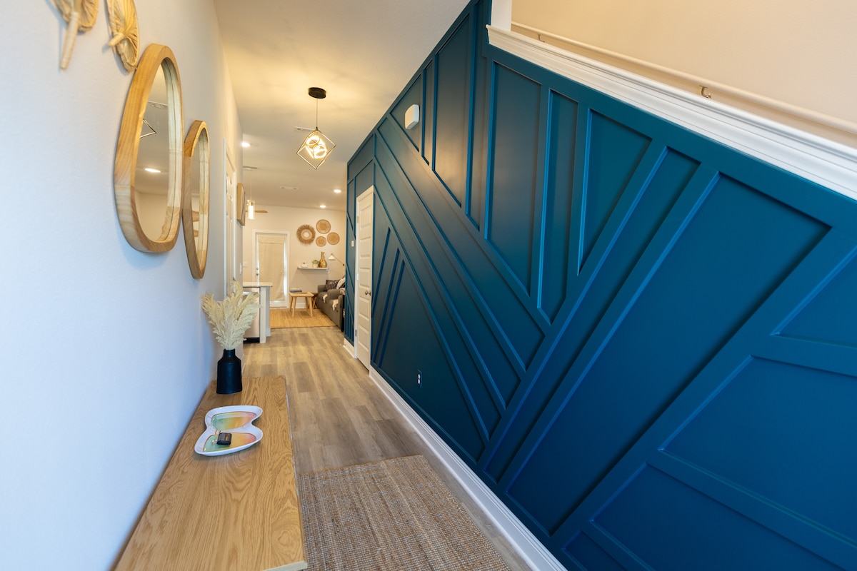 A hallway is highlighted by a bold blue feature wall, featuring geometric paneling. A wooden console table is positioned next to the wall, decorated with a small tray and a plant. Soft lighting illuminates the pathway leading to a cozy living area in the distance.