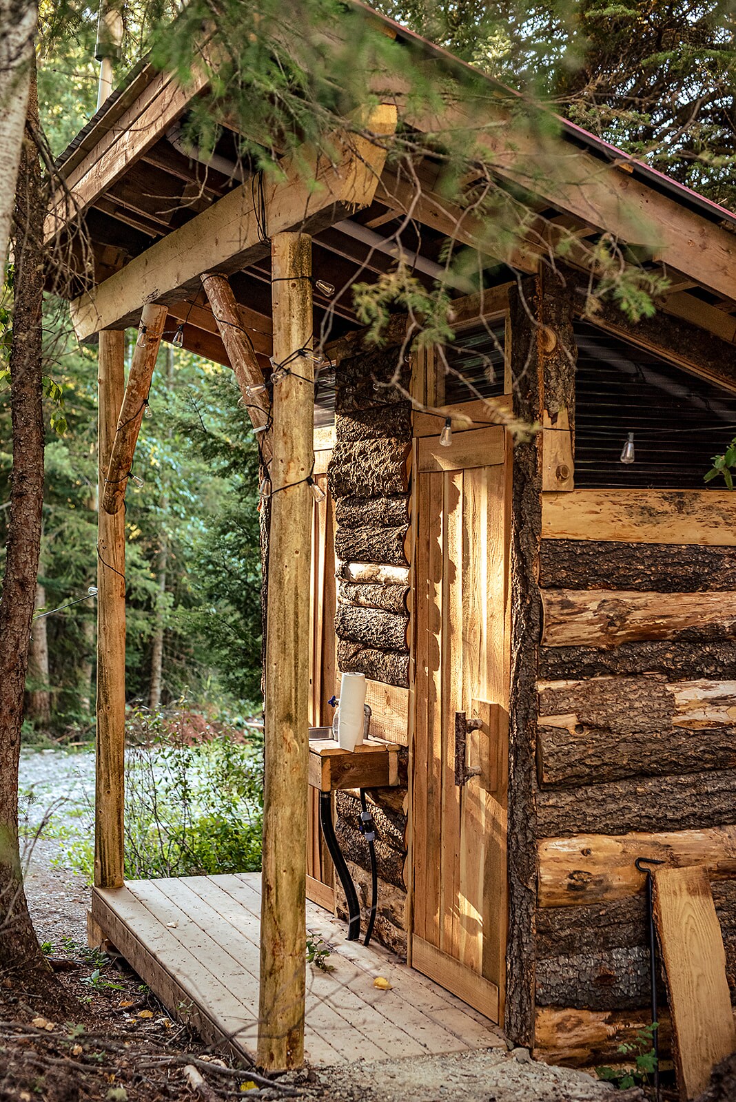 An inviting wooden entrance to a glamping unit features a sturdy door and a compact outdoor sink set on a small table. Tall trees frame the entrance, enhancing the natural setting. Soft lighting is visible above, creating a welcoming atmosphere.
