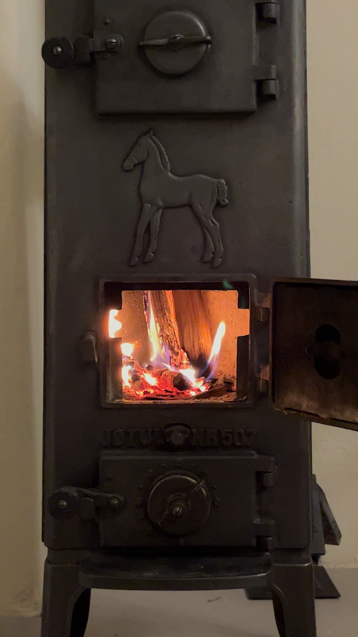 A traditional wood-burning stove stands with an illustration of a horse on the front. The door is open, revealing glowing embers and flames, providing warmth and a cozy ambiance to the room.