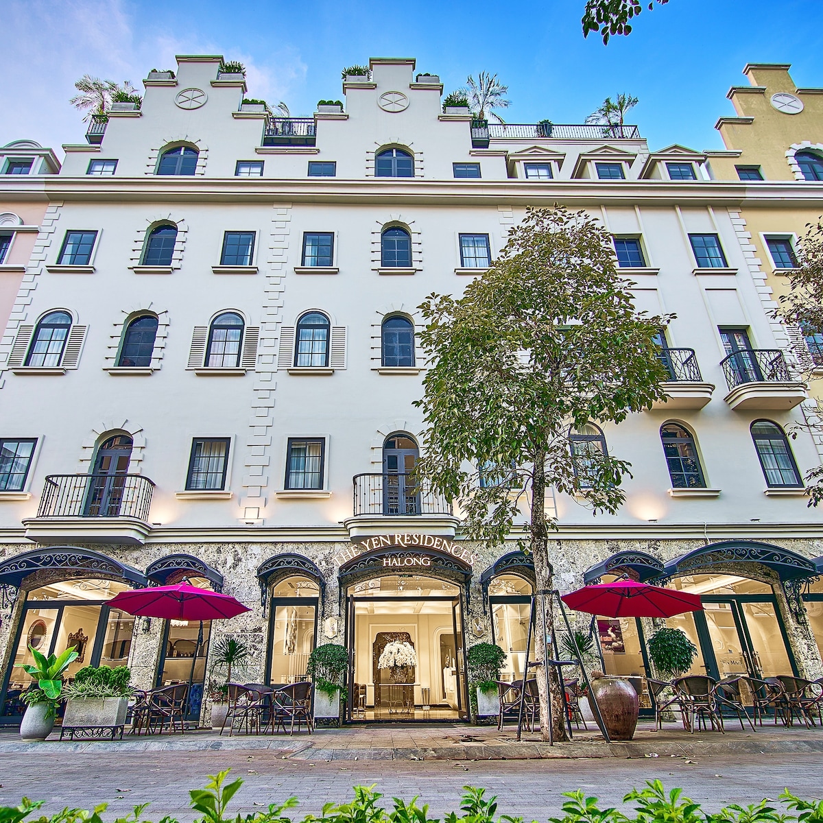 The exterior of a stylish hotel is displayed, showcasing a three-story building with ornate architectural details. Large windows are framed with wrought iron balconies. Outdoor seating with umbrellas is visible alongside potted plants, providing a welcoming entrance.