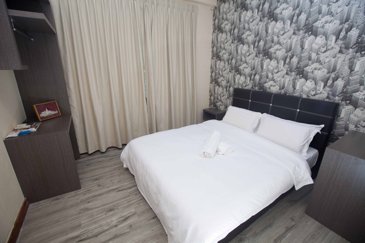 A simple and modern bedroom features a spacious bed with crisp white linens. A wall adorned with a black-and-white cityscape mural creates a focal point. Light, flowing curtains frame the window, and a minimalist wooden nightstand and dresser complement the decor.
