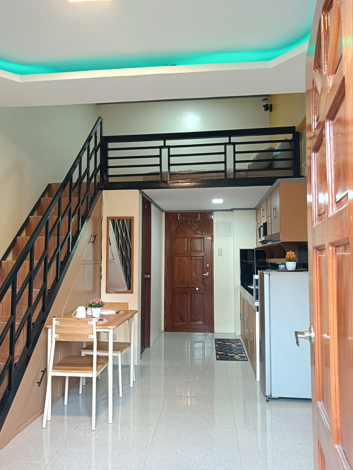 An airy living space is depicted, featuring a staircase leading to an upper level. A compact dining area with light wooden chairs is visible, along with an inviting kitchenette. Bright lights reflect off the polished floors, enhancing the open layout.