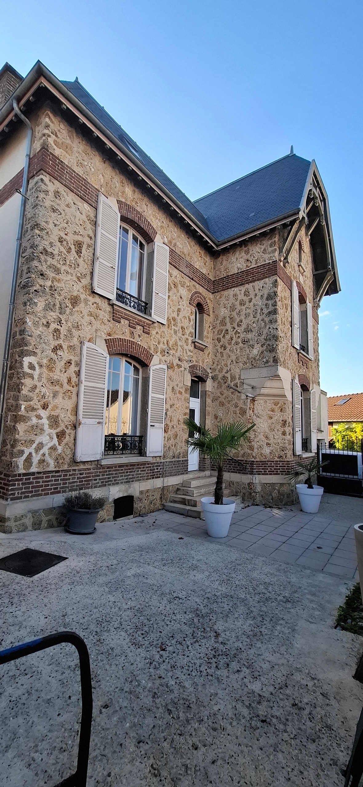 The exterior of a vintage stone house features a charming façade with large, shuttered windows. A small palm tree adds greenery to the entrance, while potted plants are positioned along the pathway. The sky above is clear, contributing to a bright atmosphere.