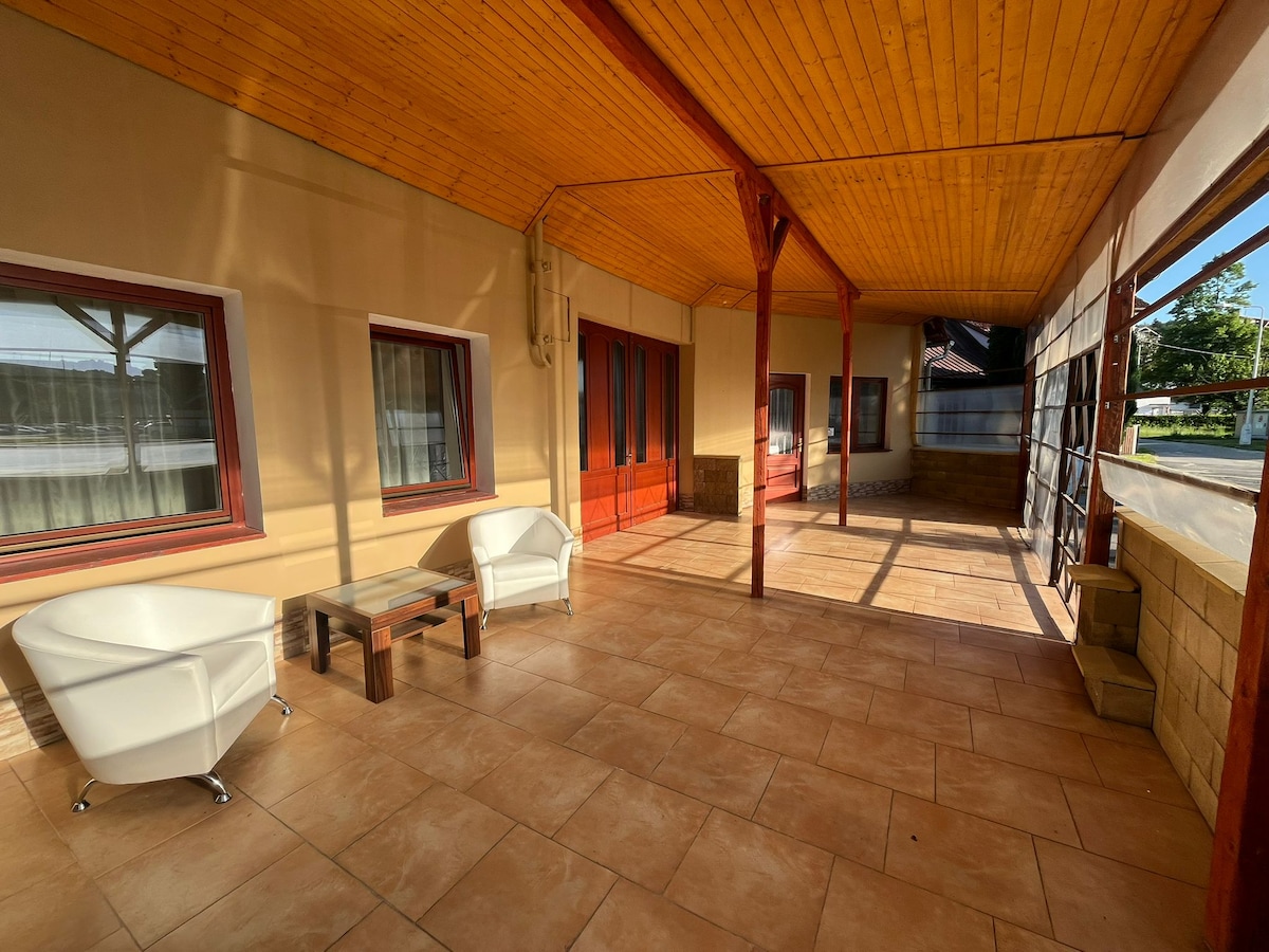An outdoor terrace area is shown with tiled flooring and wooden beams overhead. Two white chairs are positioned around a small wooden table. Large windows with red frames allow natural light to fill the space, providing a relaxed environment for gathering.