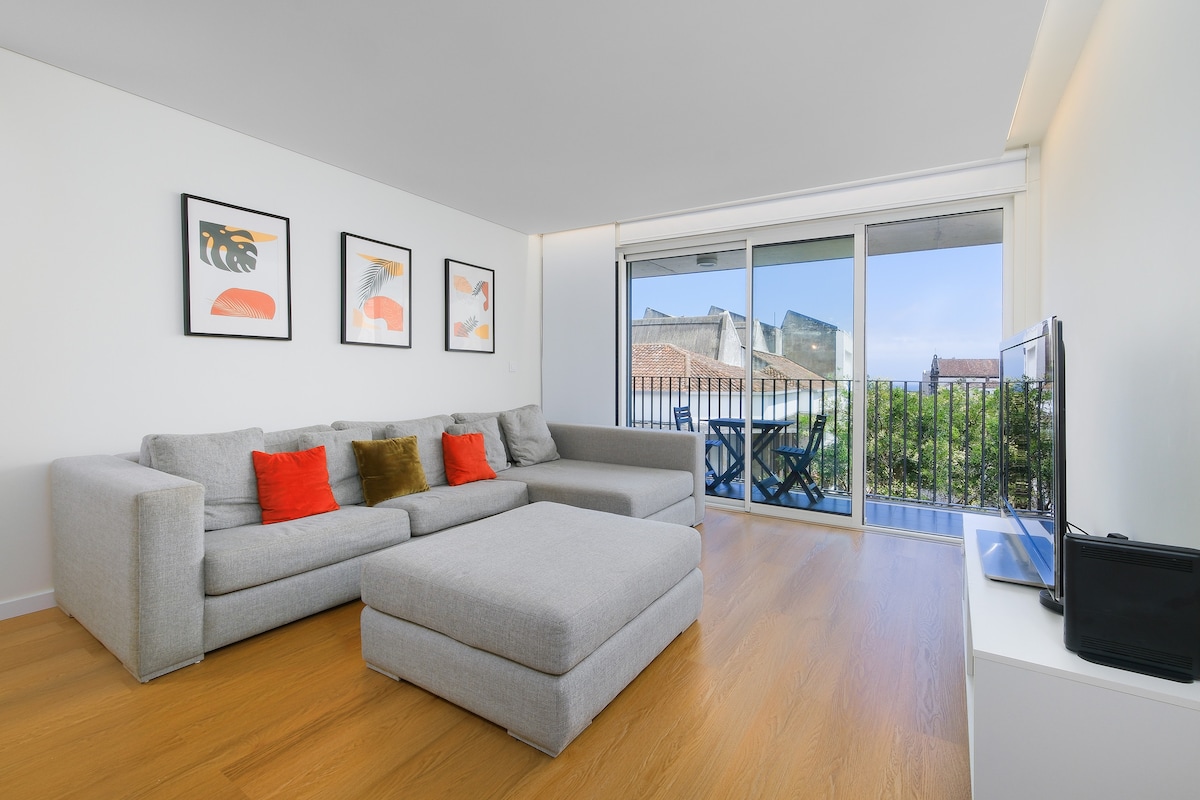 A spacious living area features a large, light grey sectional sofa with colorful accent pillows. Large windows allow natural light to brighten the room, showcasing a view of an outdoor balcony equipped with two chairs. A flat-screen television is positioned against a white wall.