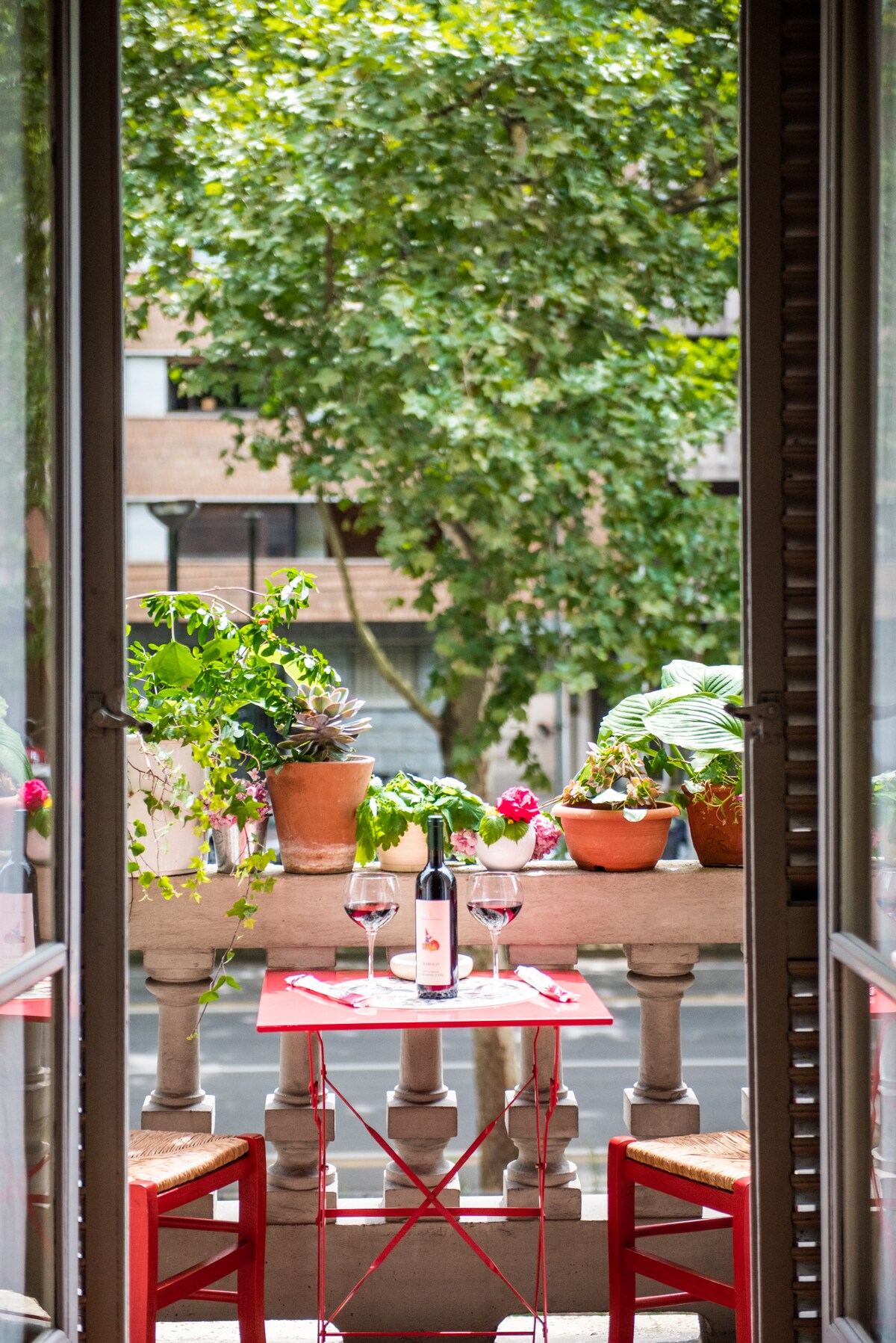 A small balcony features a red table set for two, including wine glasses and a bottle. Potted plants add greenery, while views of a tree-lined street enhance the atmosphere. The open doors provide a connection to the lively exterior.