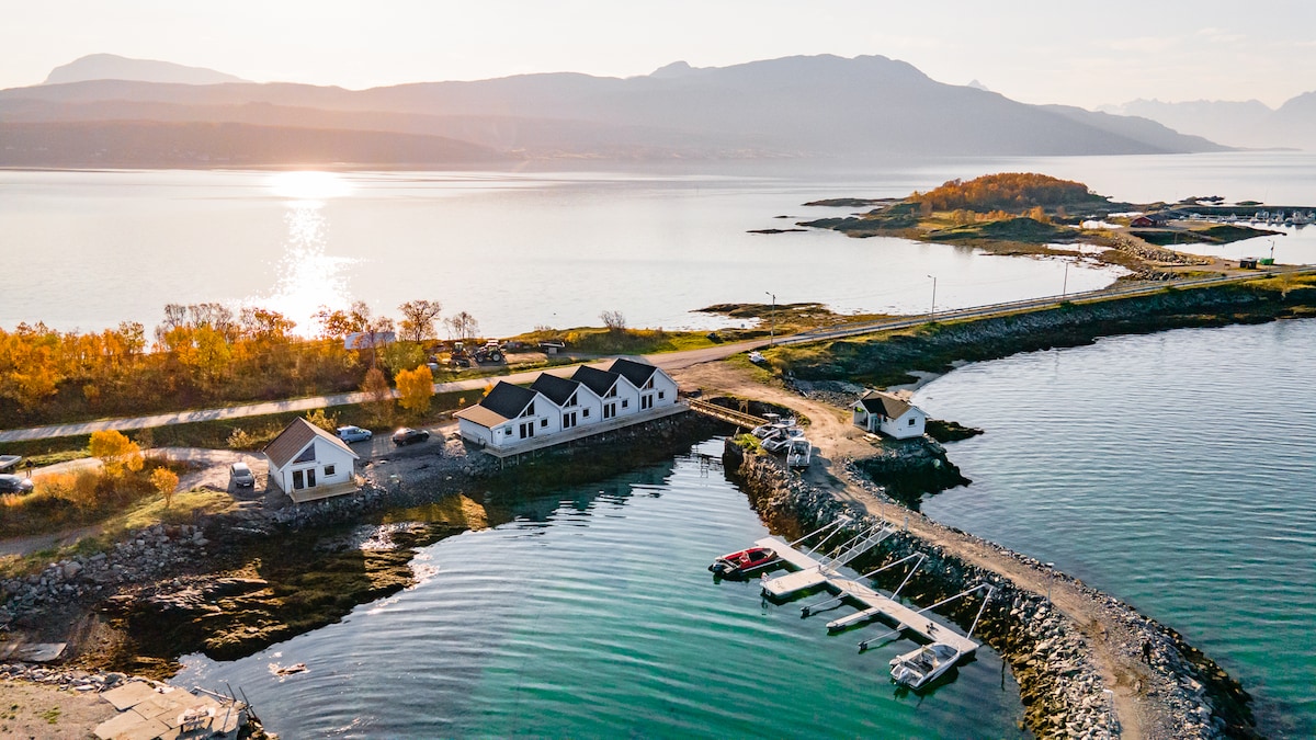 An aerial view captures modern sea lodges nestled along a tranquil shoreline. The clear water reflects the soft light of sunset, while boats are moored at a nearby dock. Autumn foliage adds warmth to the landscape, enhancing the natural beauty of the environment.