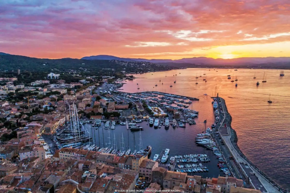 A breathtaking view captures the vibrant colors of sunset over the harbor of Saint-Tropez. The scene features numerous boats moored in the calm waters, along with hills in the background that frame the picturesque coastline.