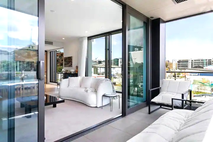 The open-plan living area is illuminated by natural light, featuring a modern white sofa and stylish armchairs. Sliding glass doors provide access to a balcony with views of the surrounding area, creating a seamless connection between indoor comfort and outdoor scenery.