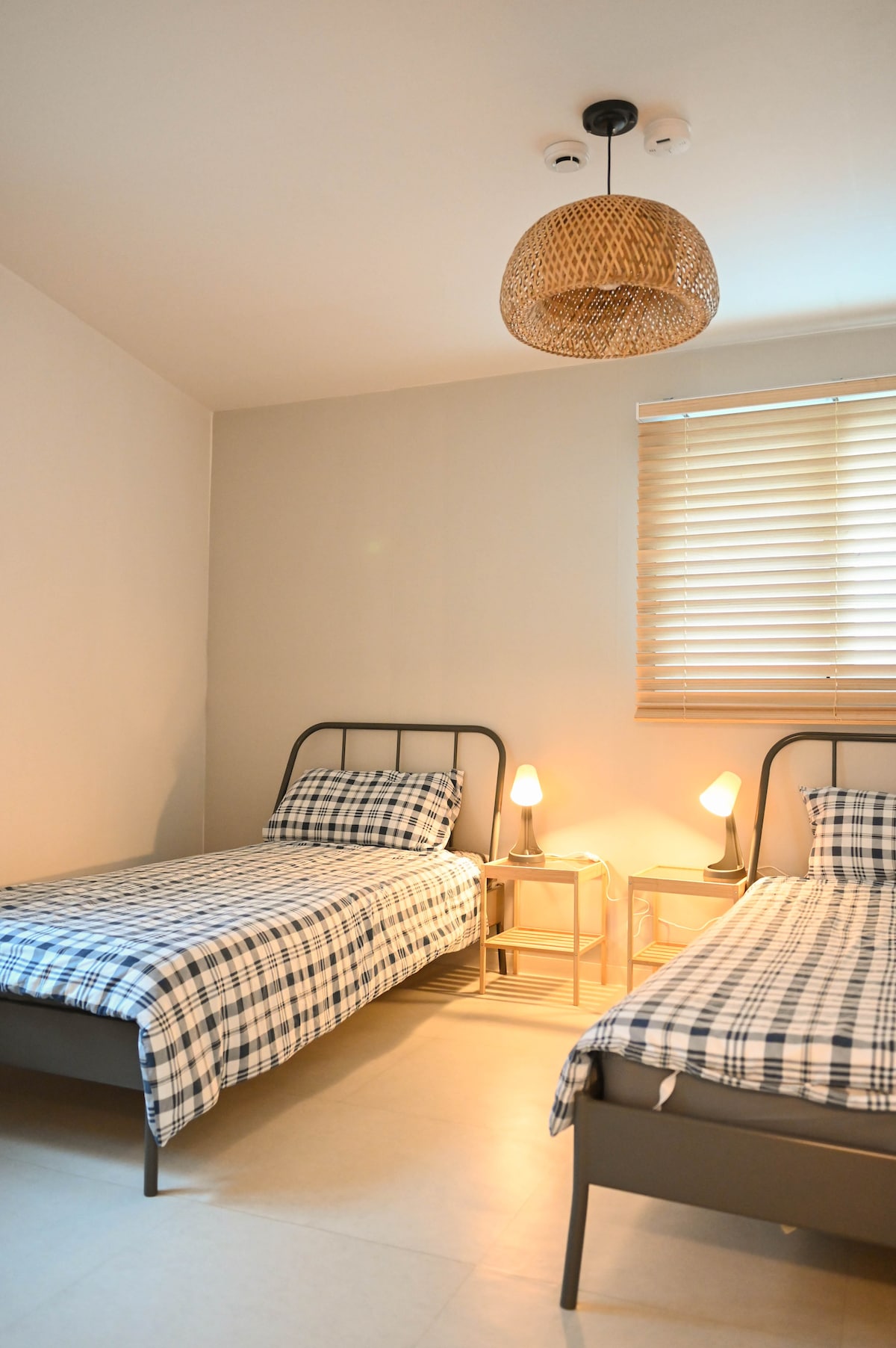 A bright room features two twin beds dressed in checkered bedding, each accompanied by a small bedside table and lamp. Natural light filters through a window with blinds, enhancing the room's serene atmosphere. A woven pendant light adds a touch of warmth.