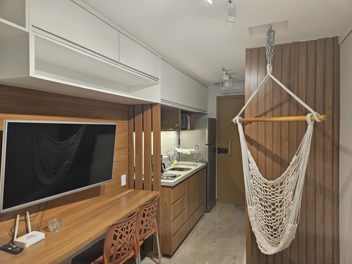 The studio features a functional kitchen with modern appliances and sleek cabinetry. A lightweight hammock hangs from a wall, while a wall-mounted TV is positioned above a wooden dining table with intricately designed chairs, creating a cozy yet practical workspace.